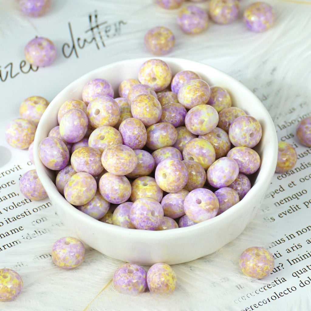 Lavender and yellow marbled round beads with holes in a small white porcelain bowl, some scattered on printed paper