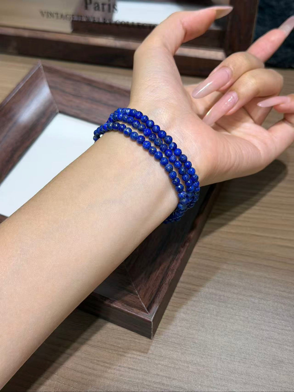 Close-up of a wrist wearing a multi-wrapped deep royal blue beaded bracelet, with long manicured nails, above a dark wooden tray.