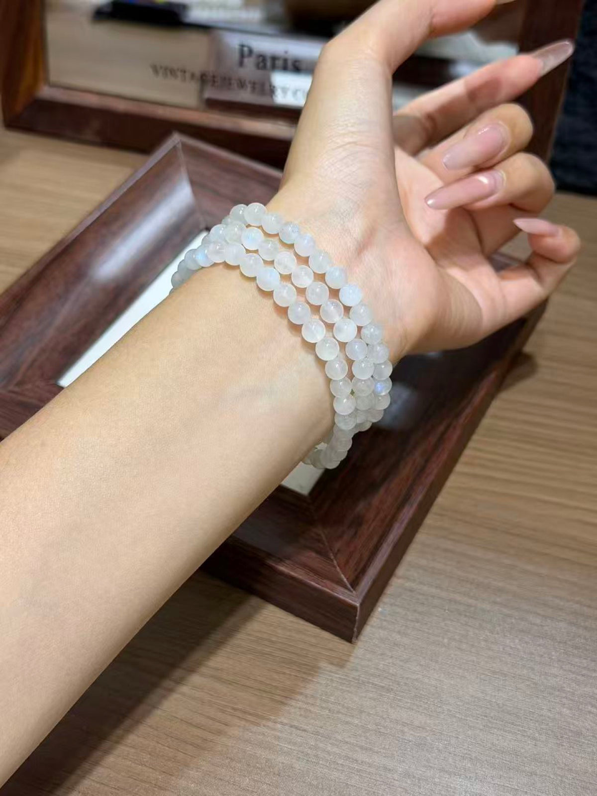 Close-up of a person's wrist wearing three strands of translucent pale blue-white round beads, resting on a dark wooden tray over a light wood tabletop