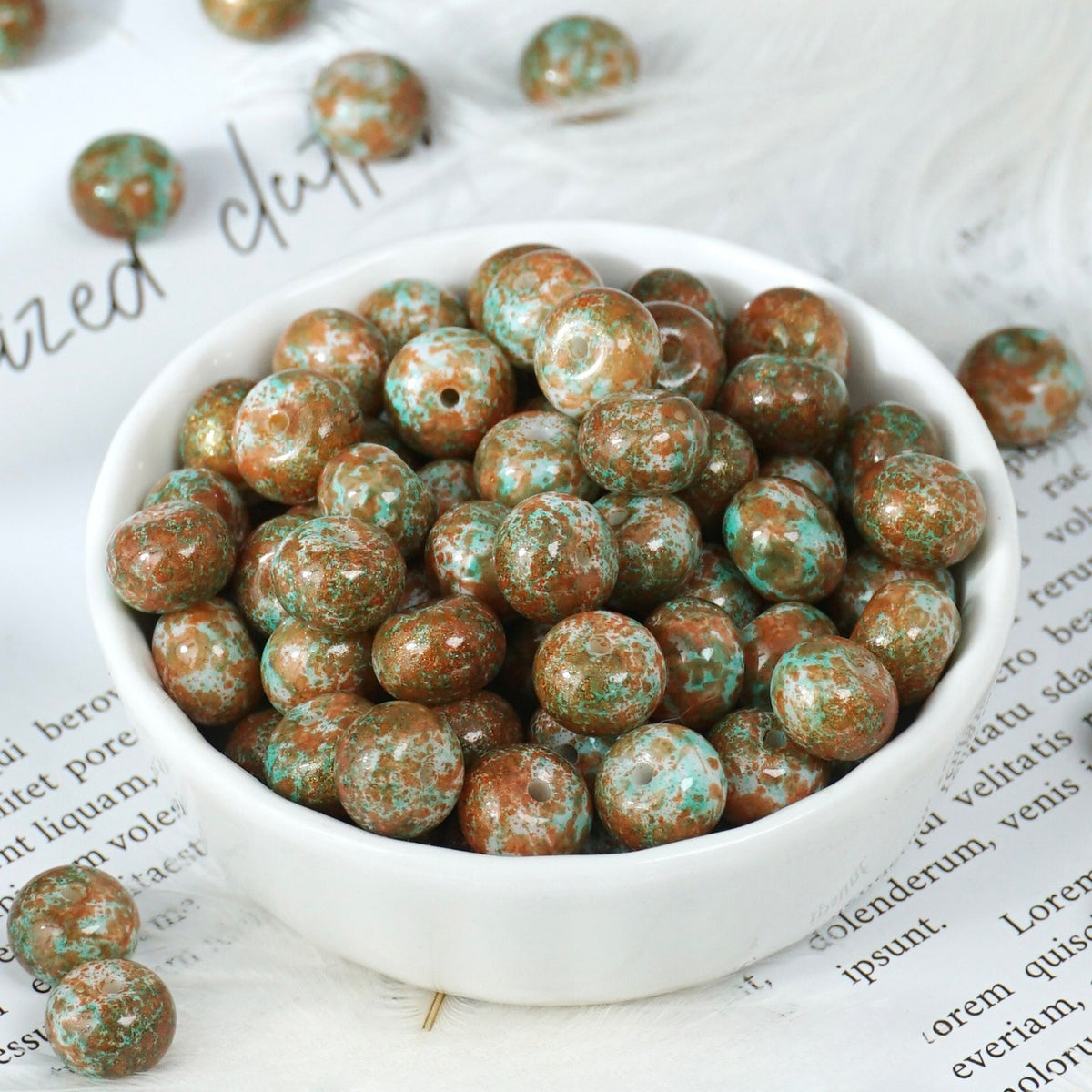White porcelain bowl of round polished beads with mottled brown, green and white stone pattern, loose beads on printed paper