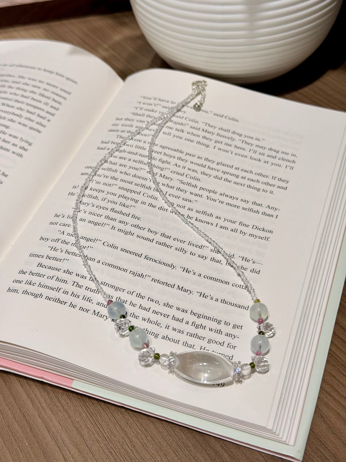 A silver-toned beaded necklace with a clear oval pendant and pale pink beads, draped over an open book on a wooden surface.