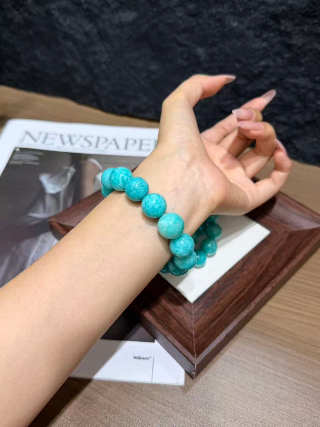 Turquoise amazonite round-bead bracelet worn on a wrist, hand resting by a wooden tray and newspaper