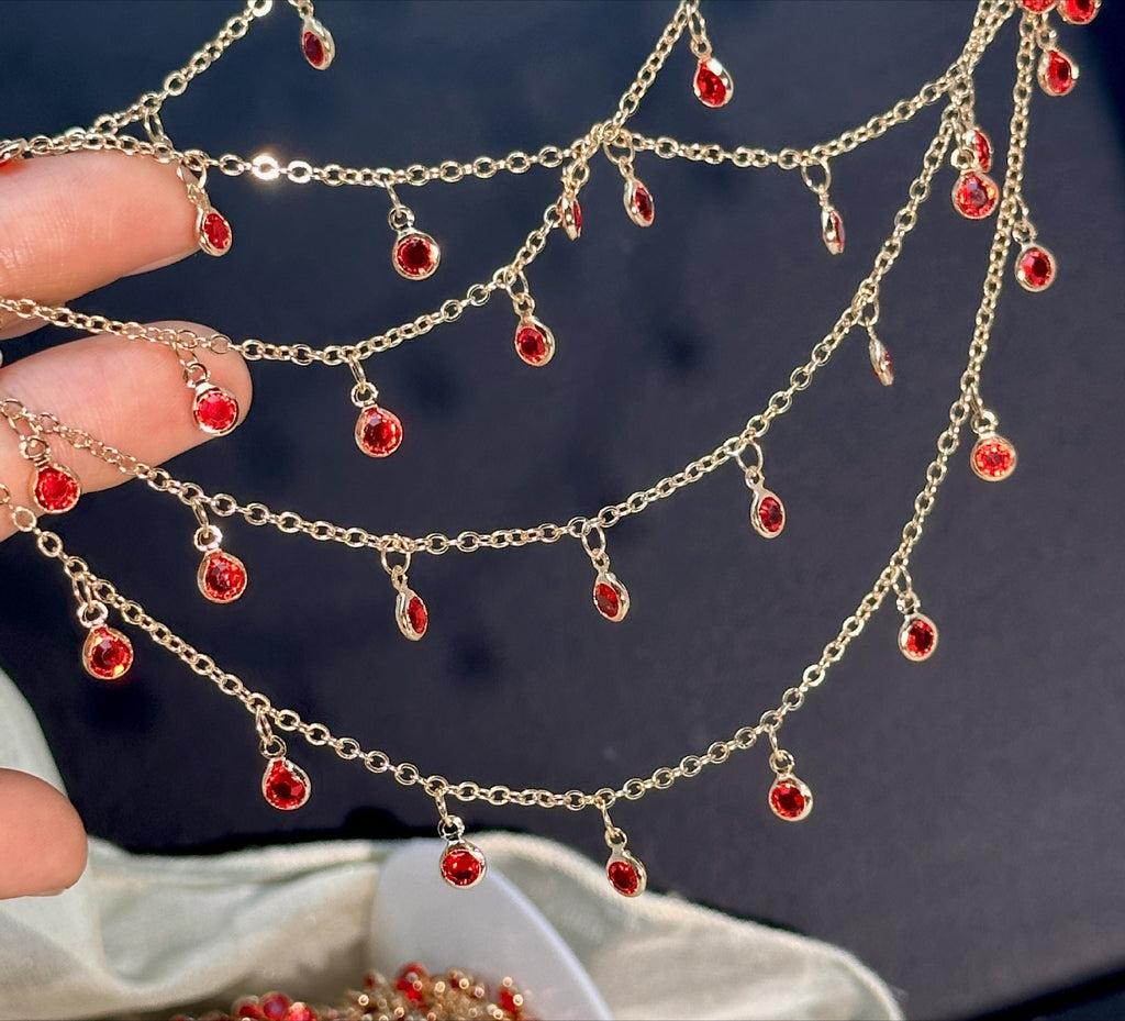 A gold-toned multi-strand chain necklace with small teardrop-shaped red gemstones, held by a hand against a dark background.