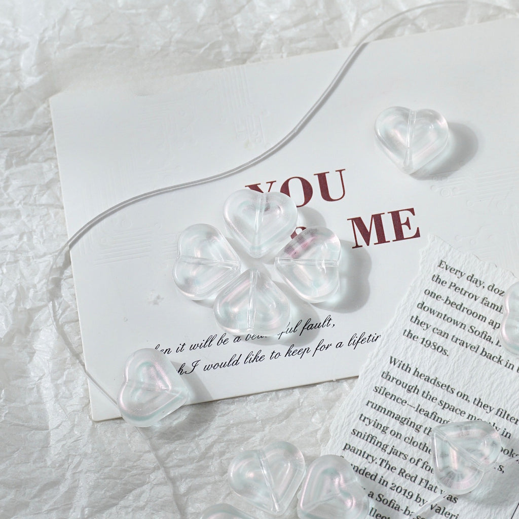 Clear heart-shaped glass beads with a four-leaf clover design, arranged on a white textured paper background next to a printed card.