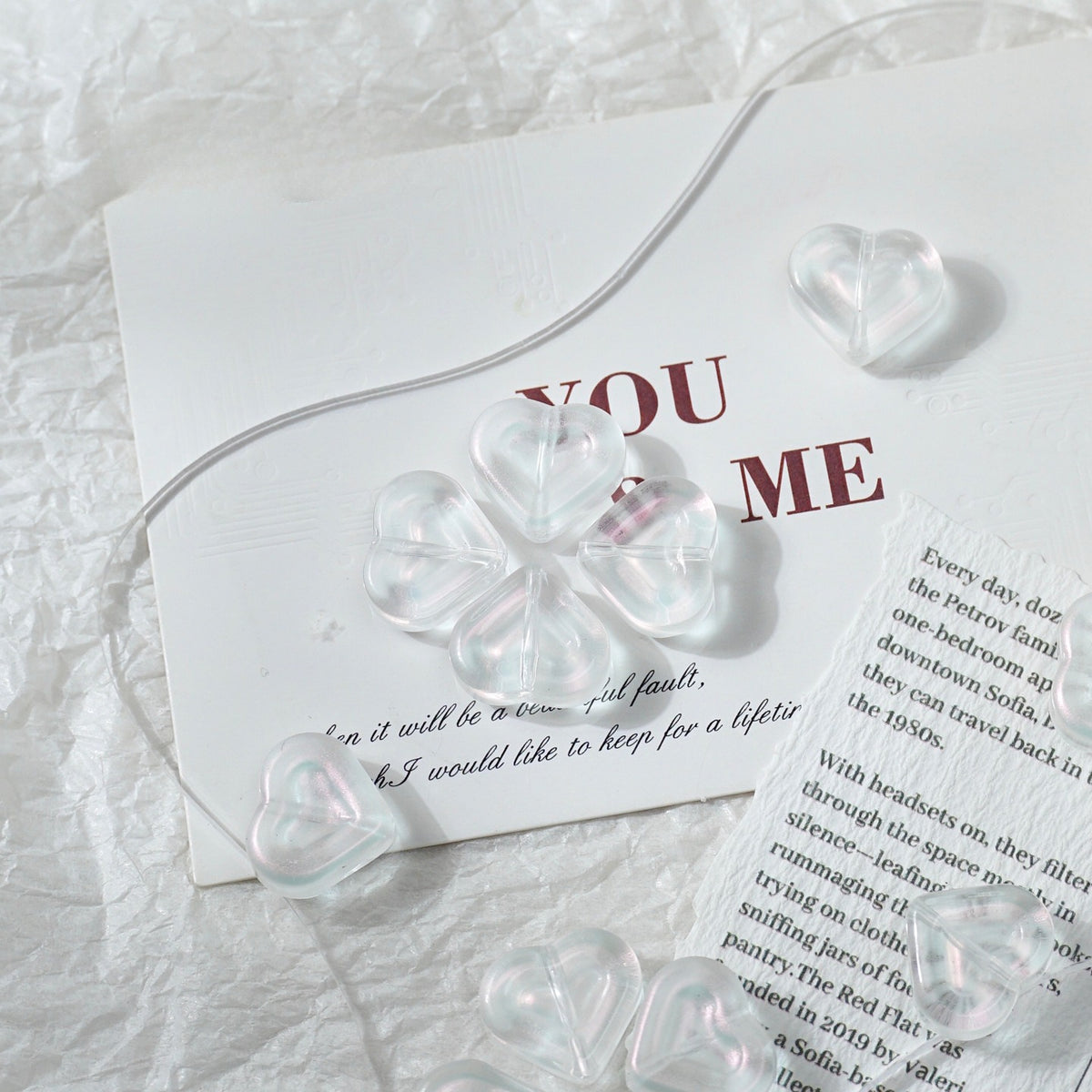 Clear heart-shaped glass beads with a four-leaf clover design, arranged on a white textured paper background next to a printed card.