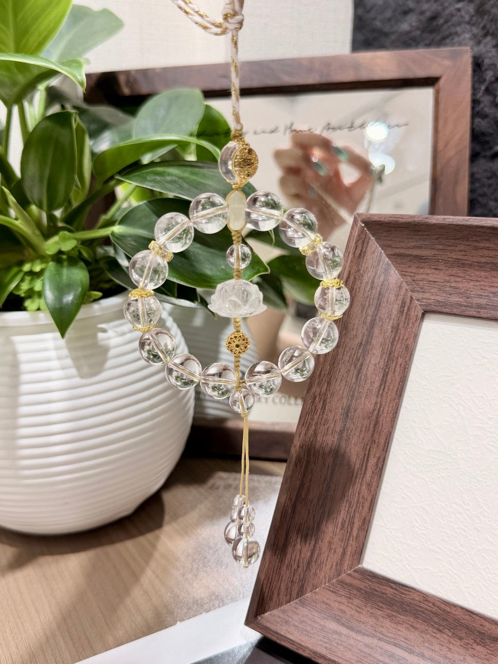 Circular clear quartz beaded car charm with carved lotus center and gold accents hanging by a potted plant
