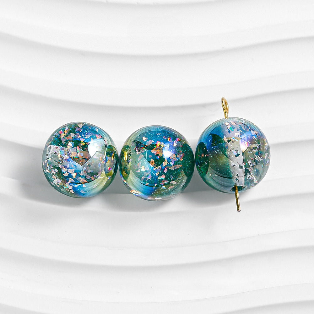 Three iridescent glitter resin beads in blue and green on a white wavy surface