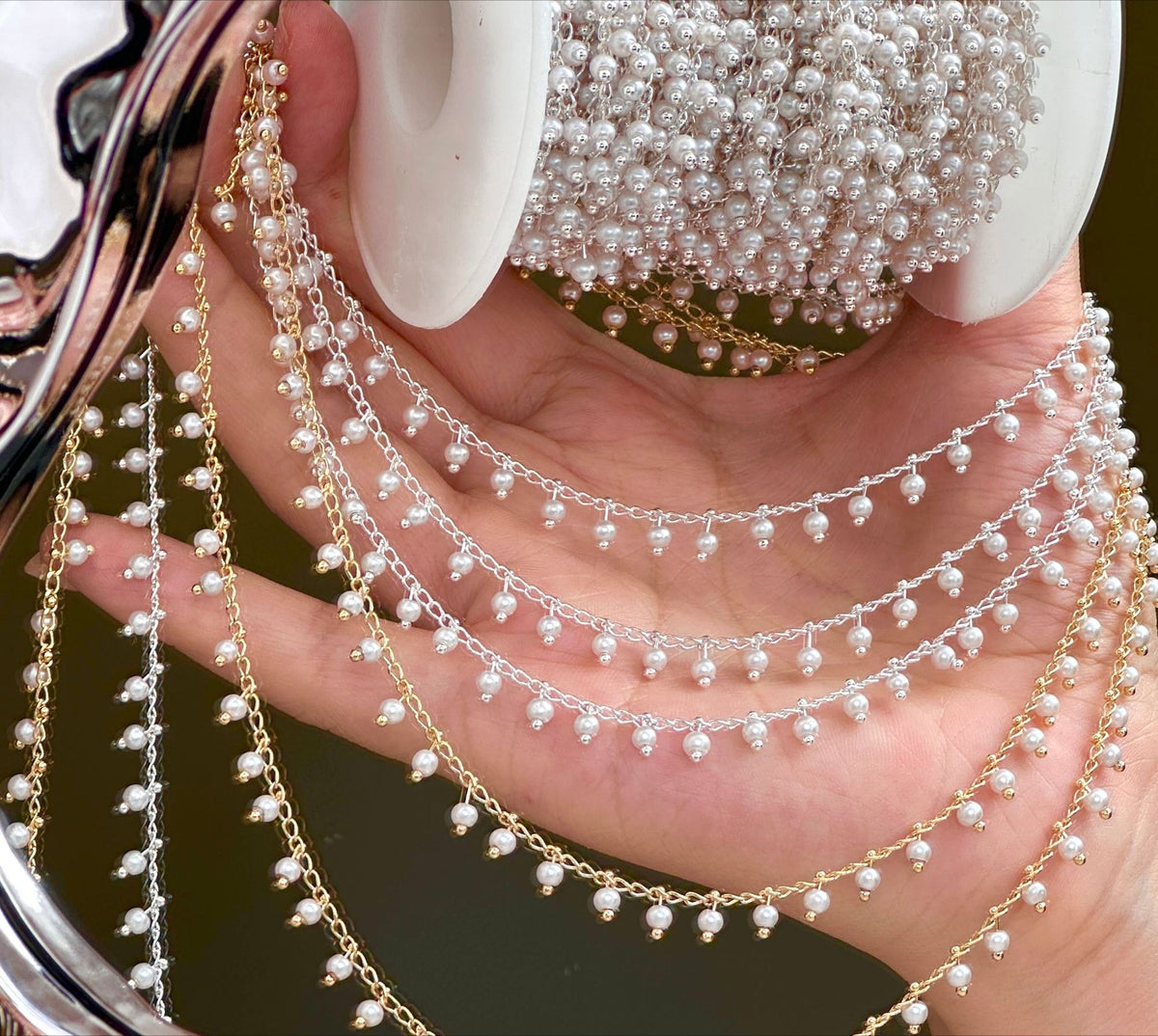 Hand holding spool of 3mm white glass pearl chains in gold and silver draped over fingers.