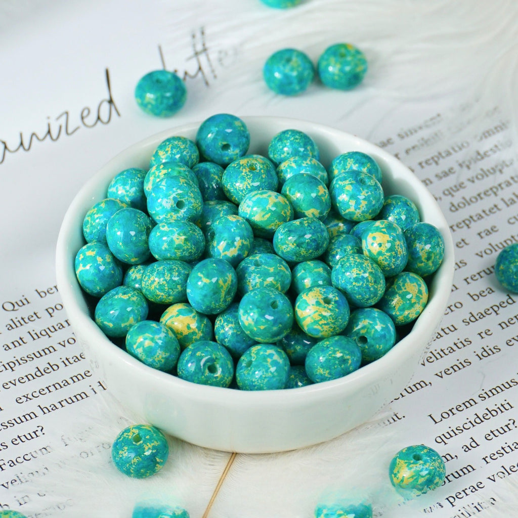 Turquoise and green marbled round beads with gold flecks in a white porcelain bowl, a few scattered on printed paper