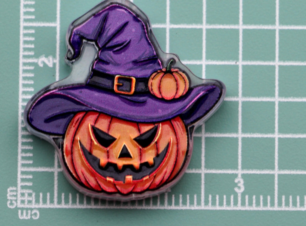 Halloween acrylic charm of an orange jack-o'-lantern pumpkin wearing a purple witch hat, on a green cutting mat