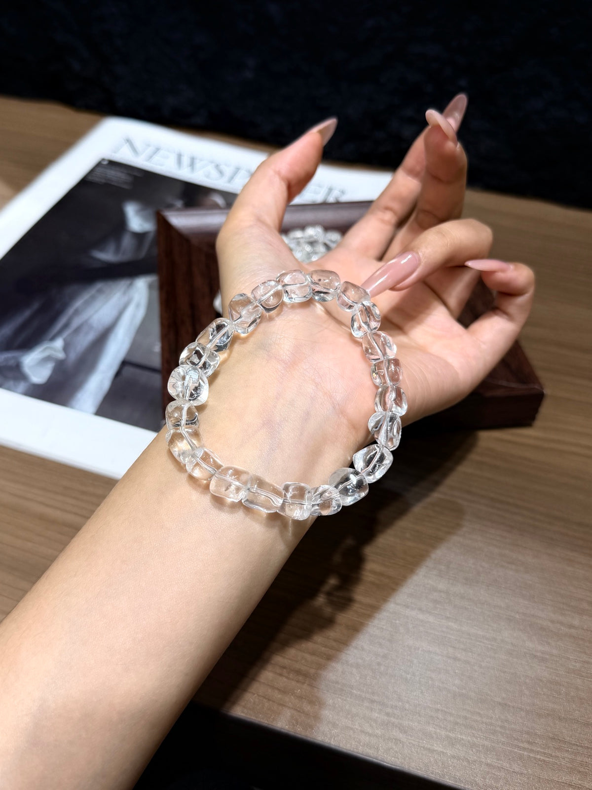 White quartz jelly-bean nugget bracelet worn on a wrist, hand posed palm-up beside a newspaper and wooden box