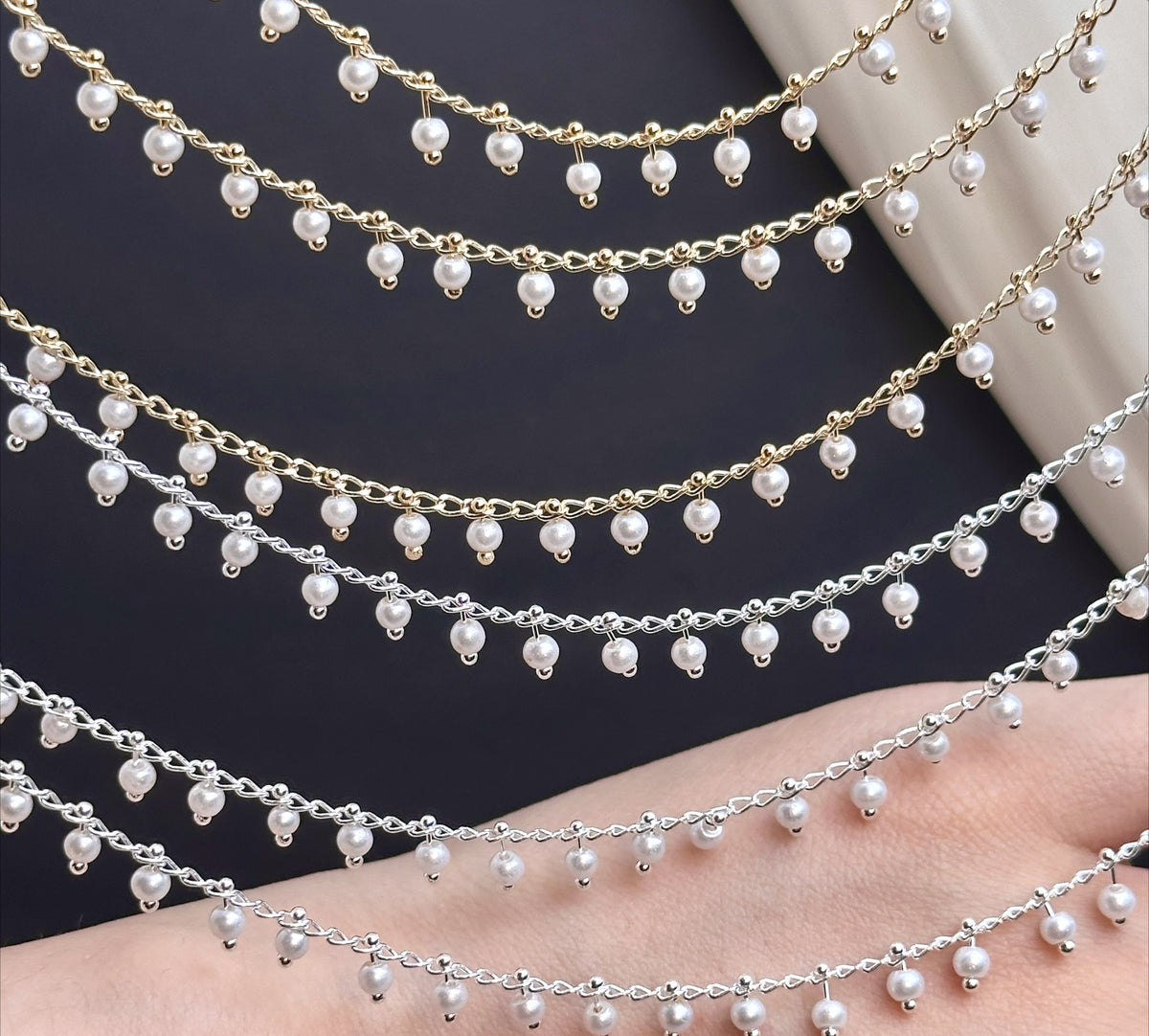 Three 3mm white glass pearl chain strands, two gold and one silver, draped across hand on dark background.