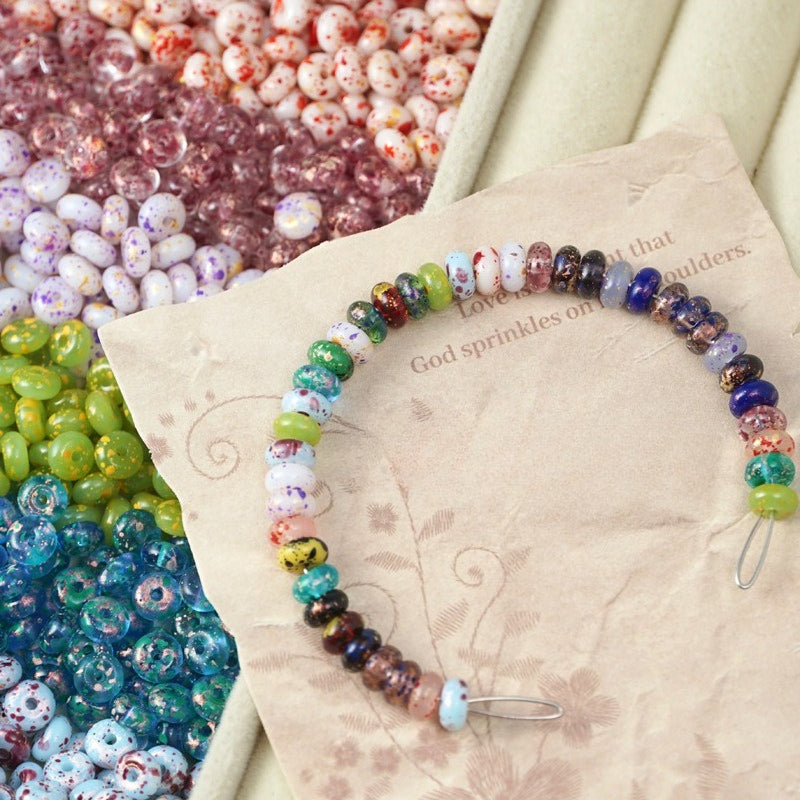 Colorful patterned glass abacus bead bracelet on beige printed card with loose green, pink, blue, white beads behind.