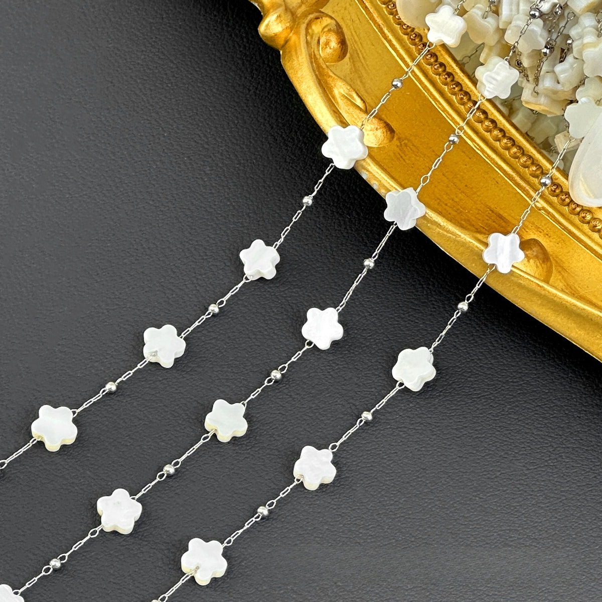 Silver station chain with white flower-shaped beads draped on dark surface beside ornate gold tray.