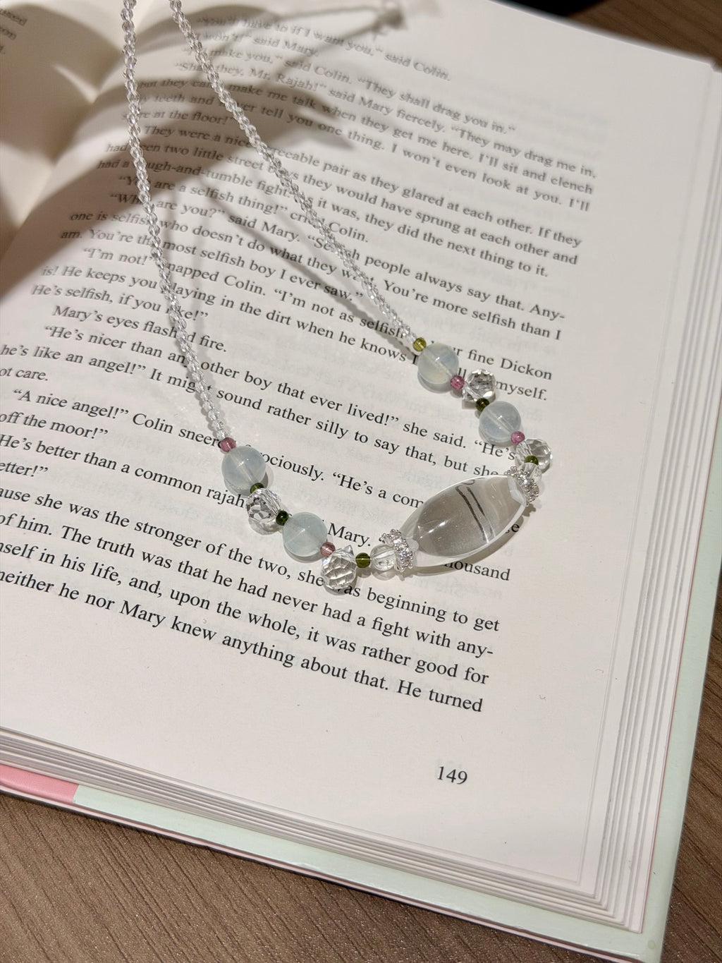 A silver beaded necklace with clear and white beads and small green accents, draped over an open book page.