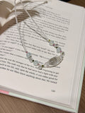 A silver beaded necklace with clear and white beads and small green accents, draped over an open book page.