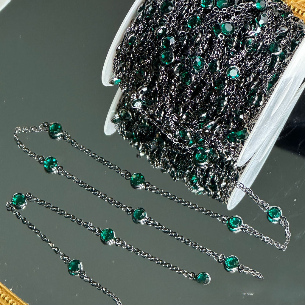 A spool of dark silver chain embellished with small round green rhinestones, with several loose chain sections displayed on a reflective gray surface.
