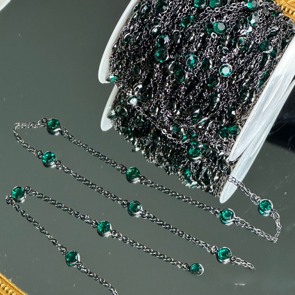 A spool of dark silver chain embellished with small round green rhinestones, with several loose chain sections displayed on a reflective gray surface.