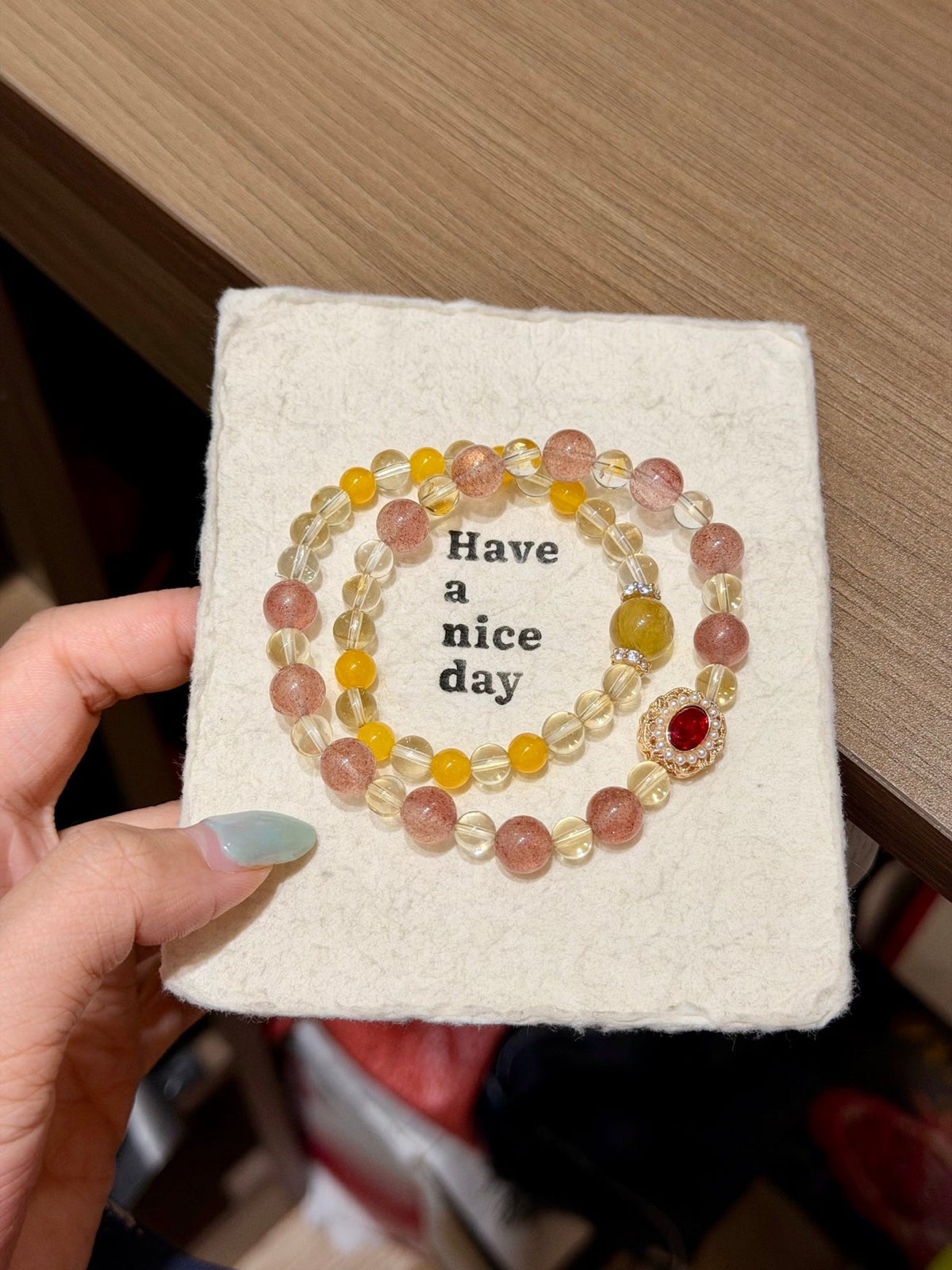 Hand holding a cream felt card printed 'Have a nice day' with a pink and yellow beaded bracelet with red gemstone