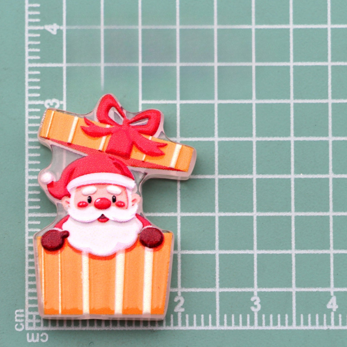 Santa peeking from orange gift box acrylic charm with red bow on green cutting mat