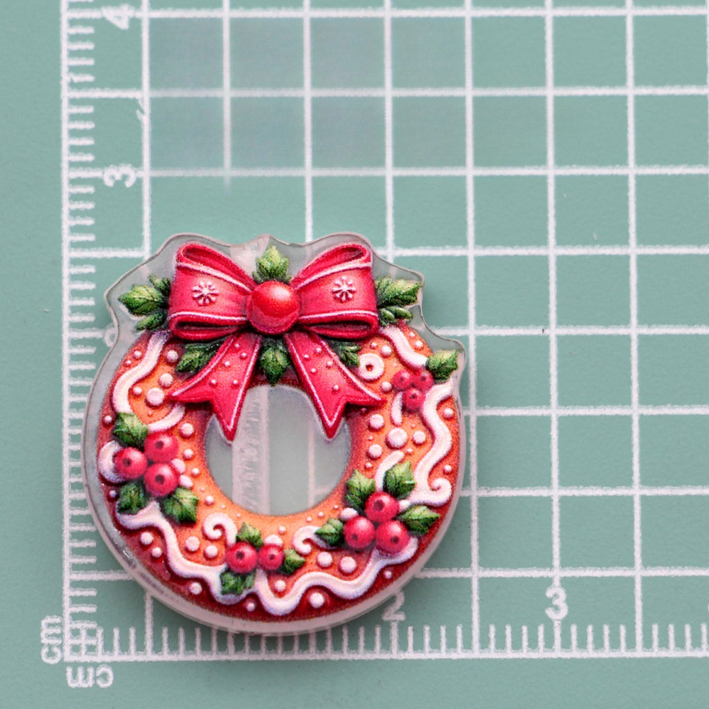 Christmas wreath acrylic charm with red bow, holly leaves and berries on green gridded mat