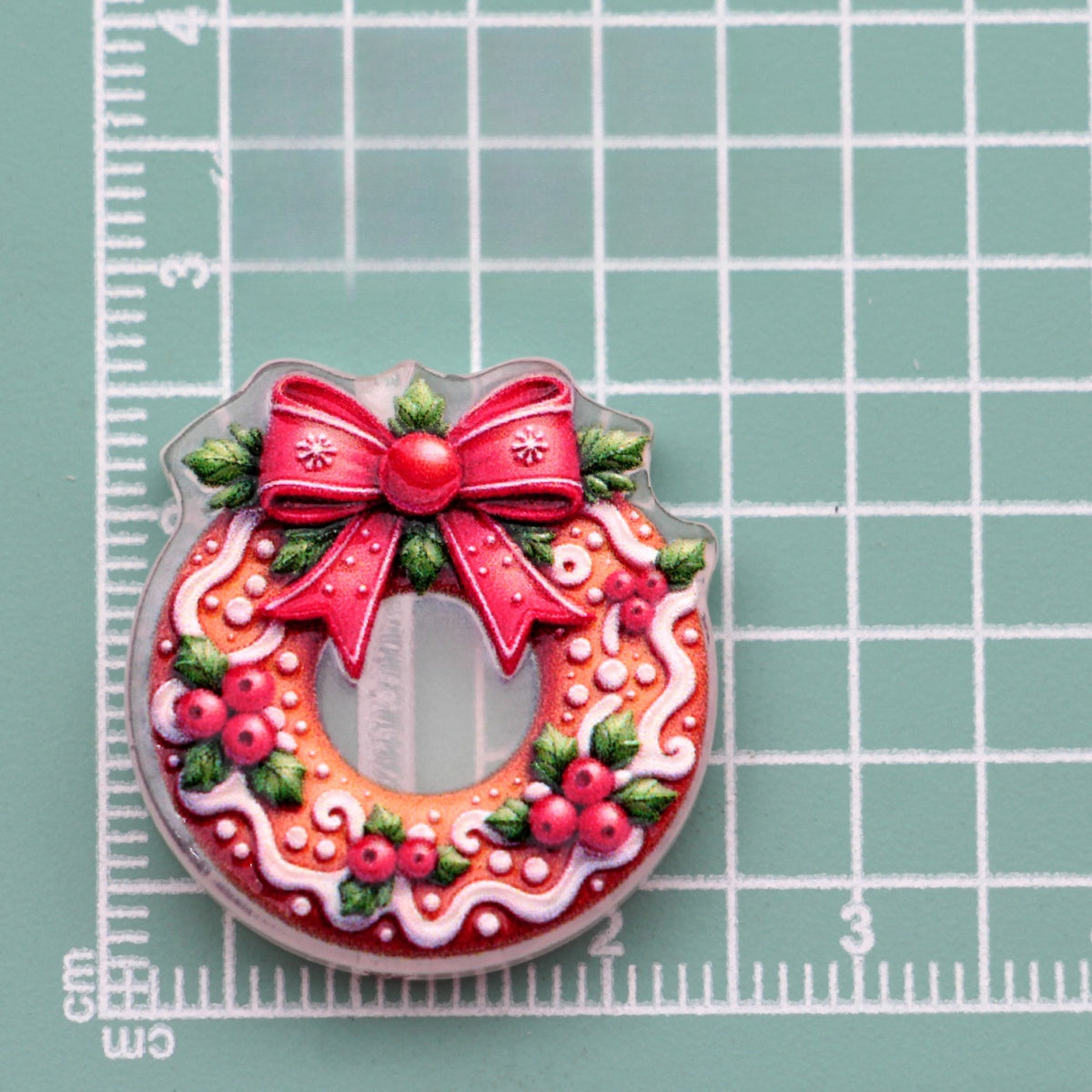 Christmas wreath acrylic charm with red bow, holly leaves and berries on green gridded mat