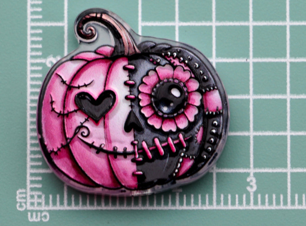 Pink and black pumpkin-shaped acrylic charm with sugar-skull design on green cutting mat with centimeter markings