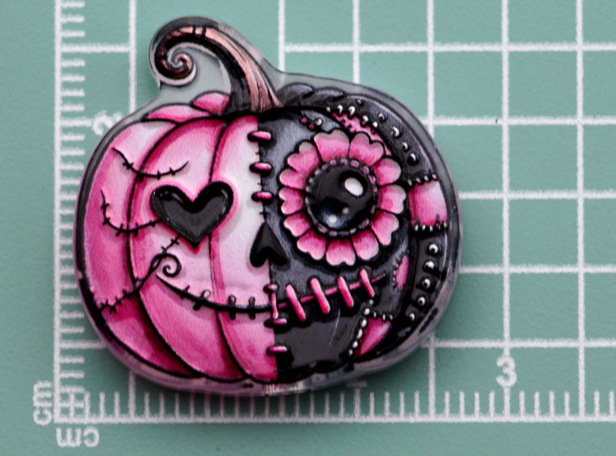 Pink and black pumpkin-shaped acrylic charm with sugar-skull design on green cutting mat with centimeter markings