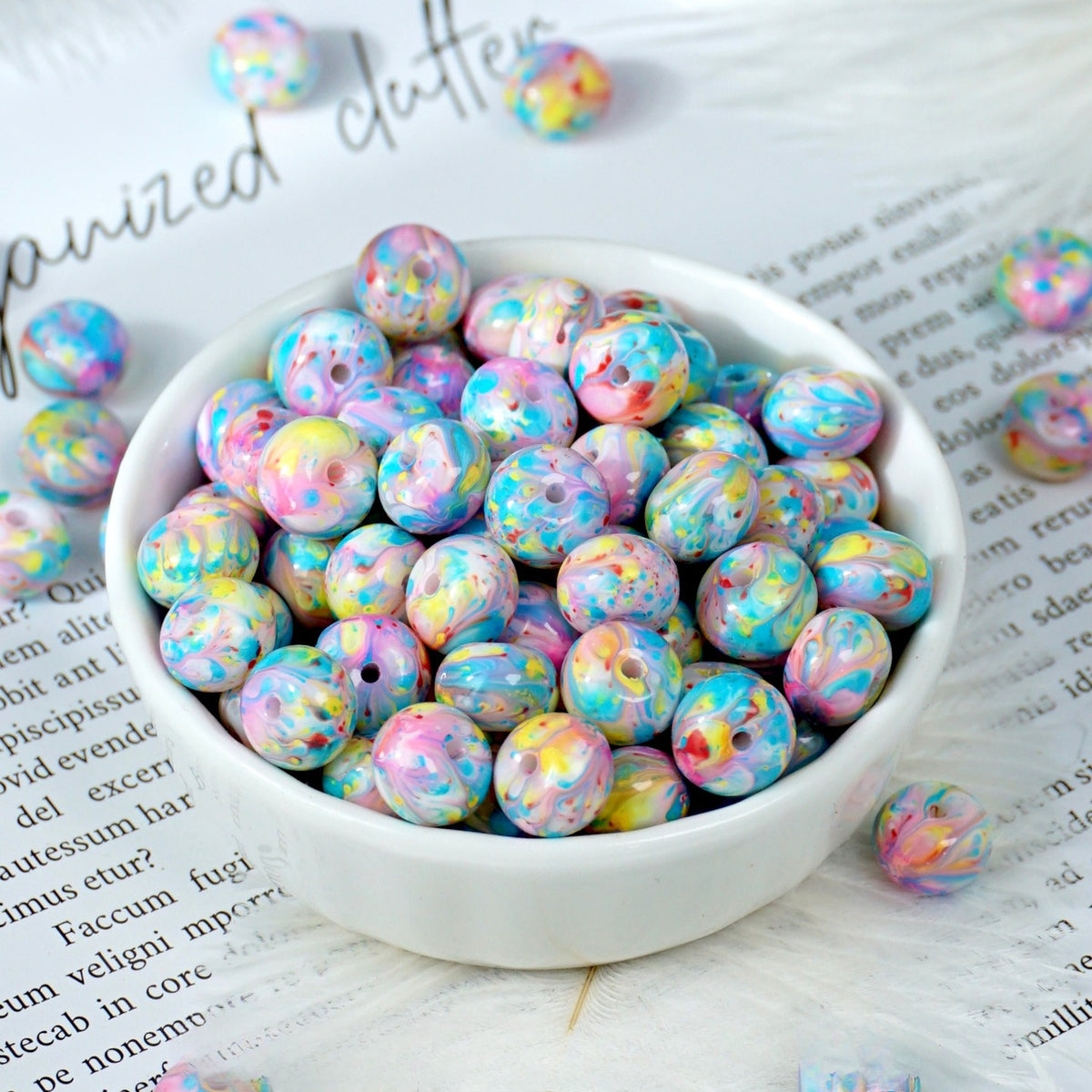Colorful marbled pastel round beads in pink, blue, yellow and purple in a white porcelain bowl, a few loose on printed paper