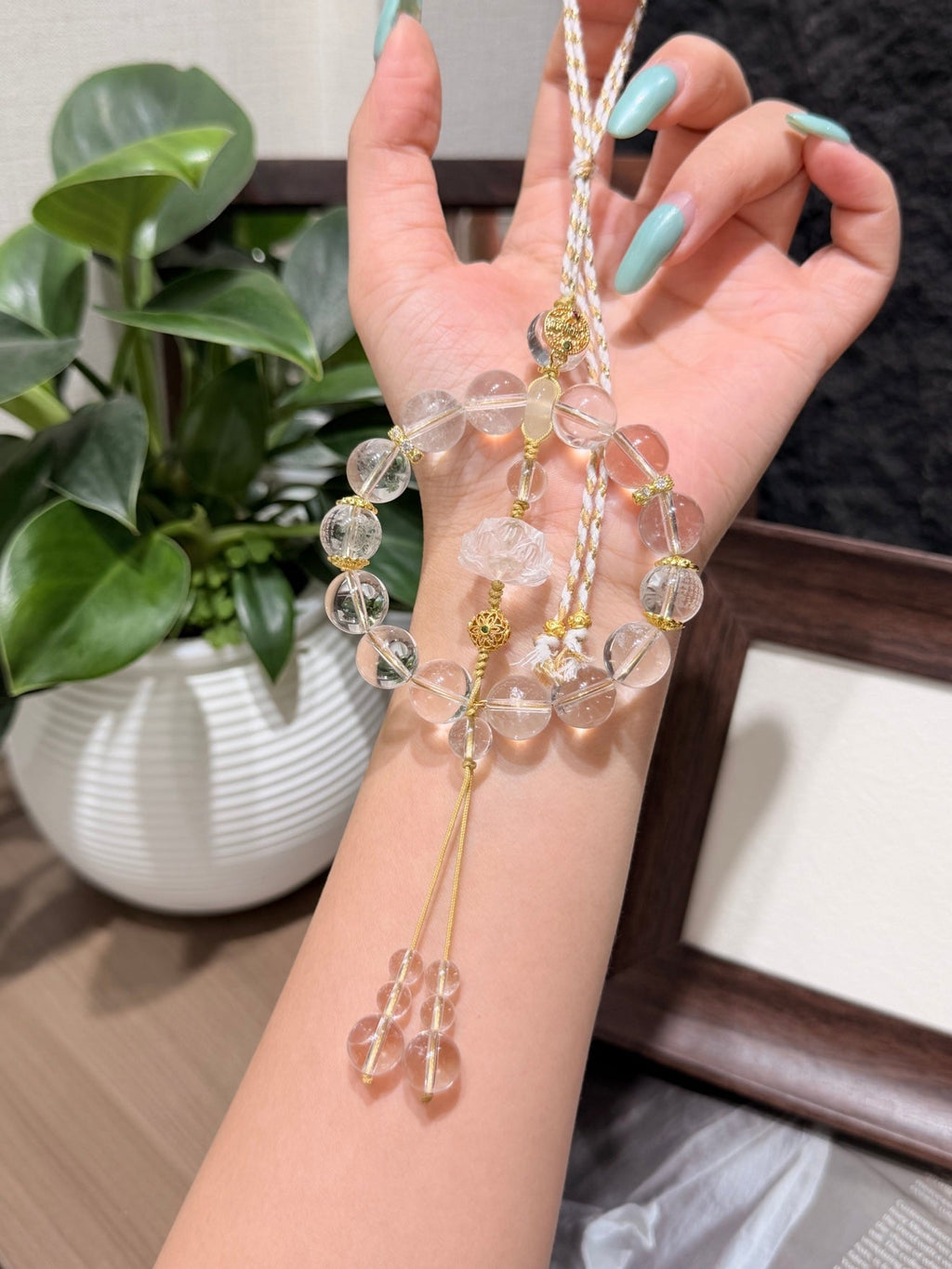 Hand holding clear quartz beaded car charm with gold spacers, champagne cord and beaded tassel, potted plant behind