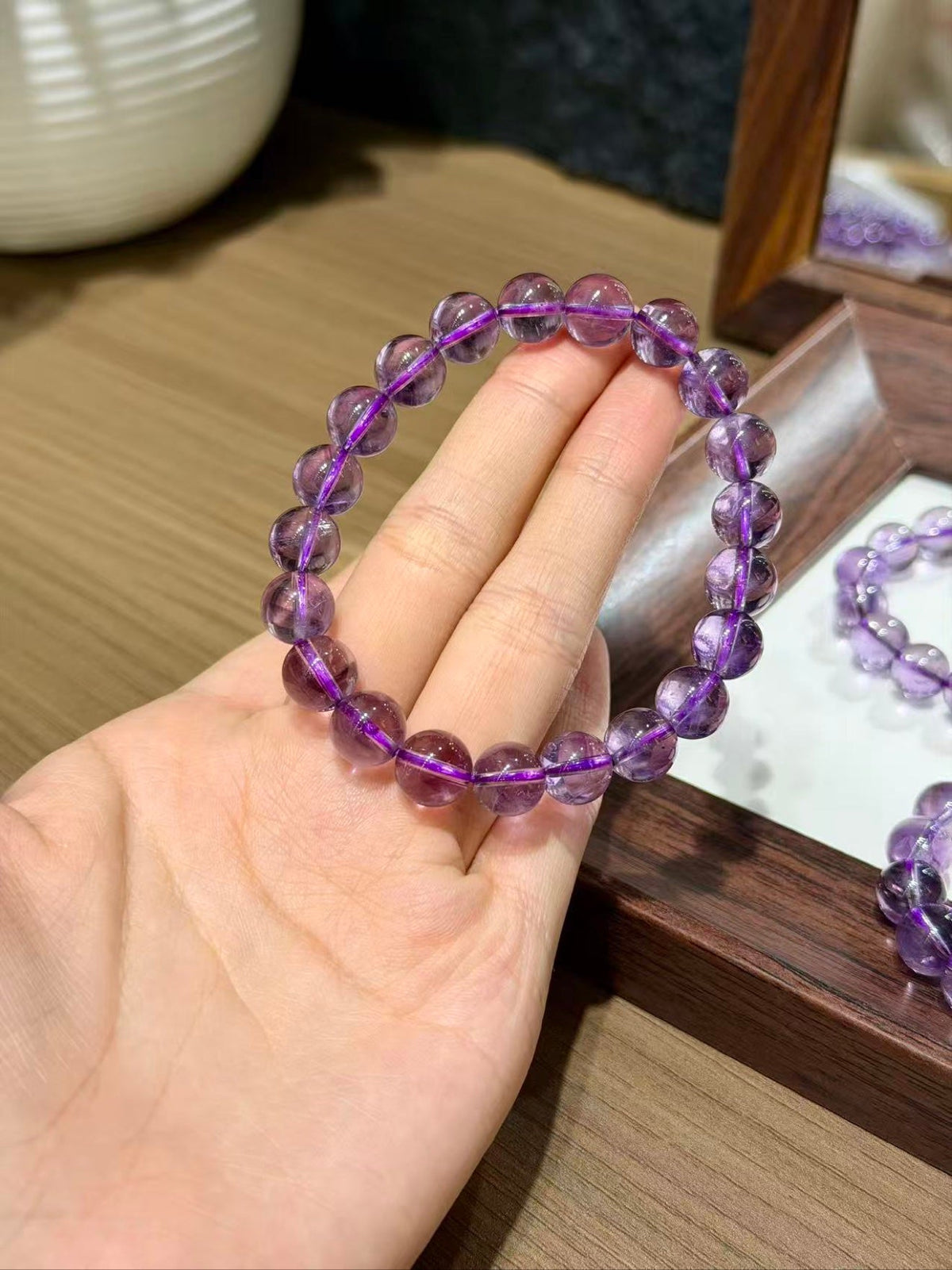 Hand holding a purple translucent amethyst beaded bracelet, wooden tray with more bracelets behind