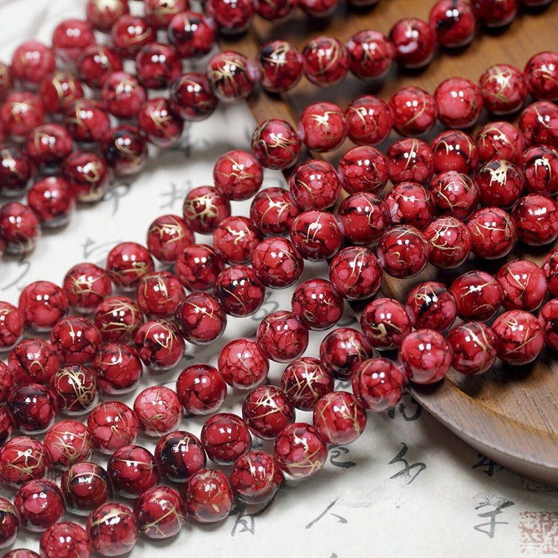Red porcelain beads with gold foil threads, arranged in strands on a light surface with a wooden tray.