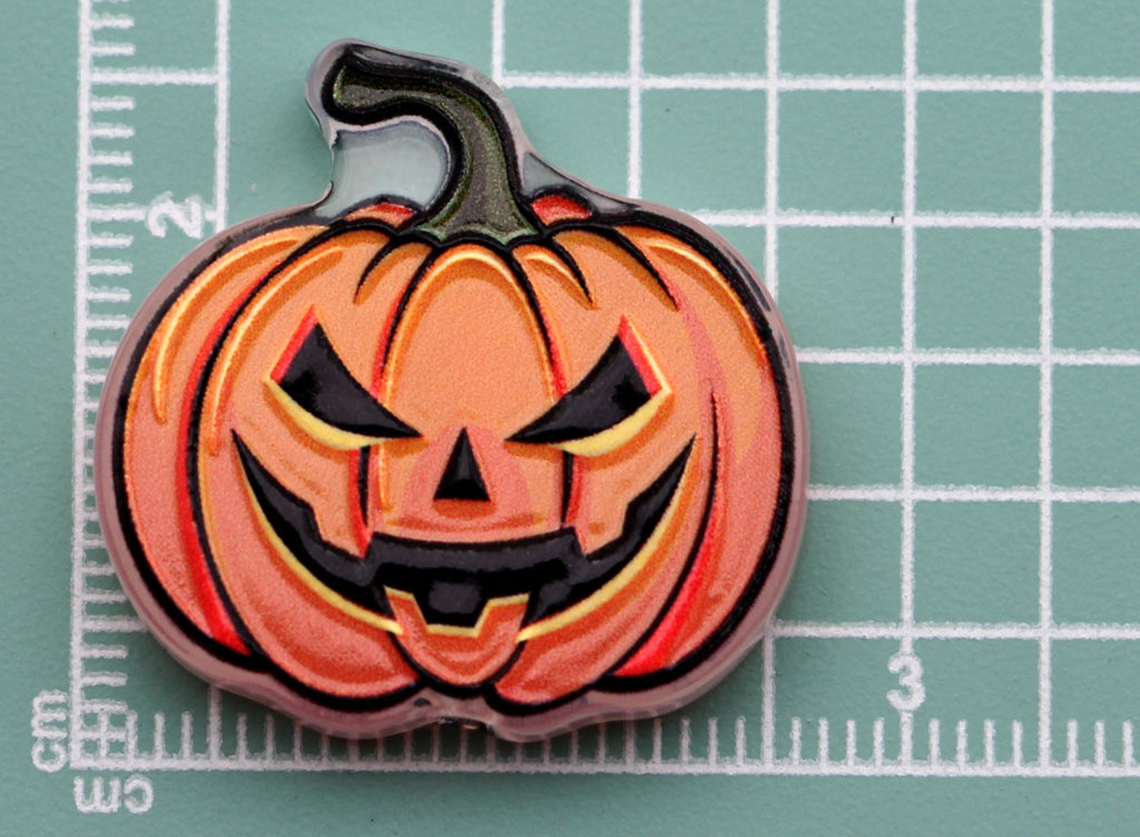 Pumpkin-shaped acrylic charm with a carved jack-o'-lantern face on a green cutting mat with grid lines