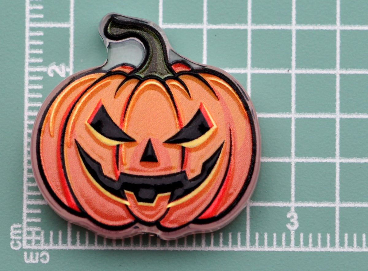 Pumpkin-shaped acrylic charm with a carved jack-o'-lantern face on a green cutting mat with grid lines