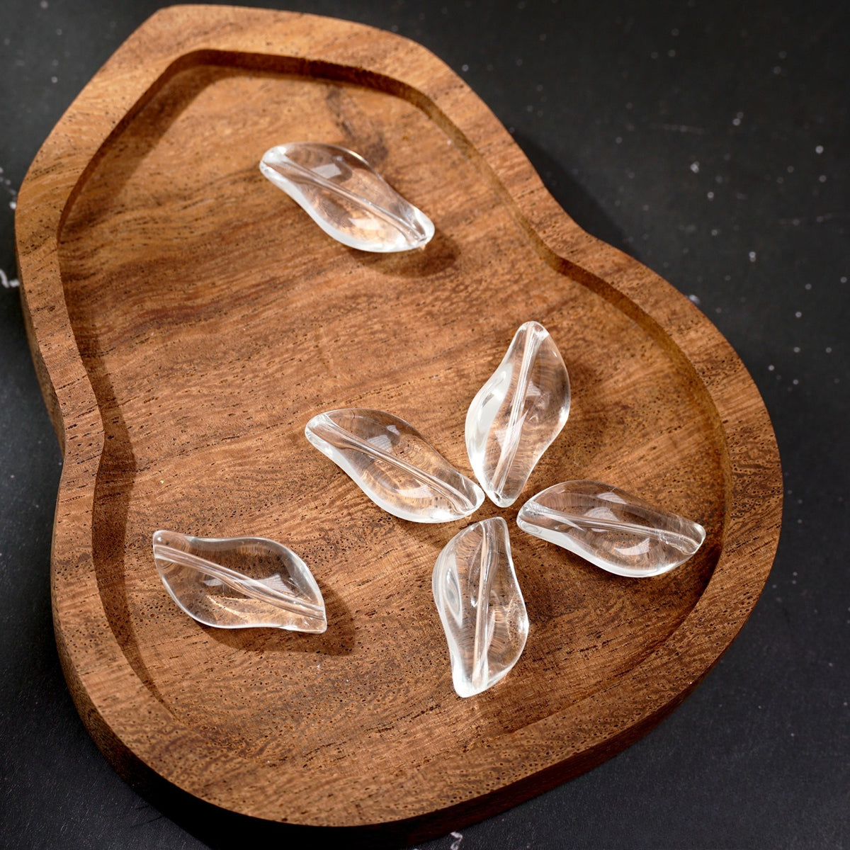 Five clear leaf-shaped glass beads on a carved wooden tray against a dark background