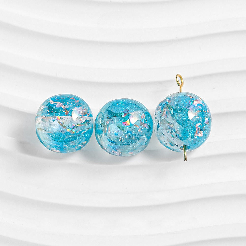 Three blue glitter resin beads with iridescent flakes on white; right bead has gold loop and pin