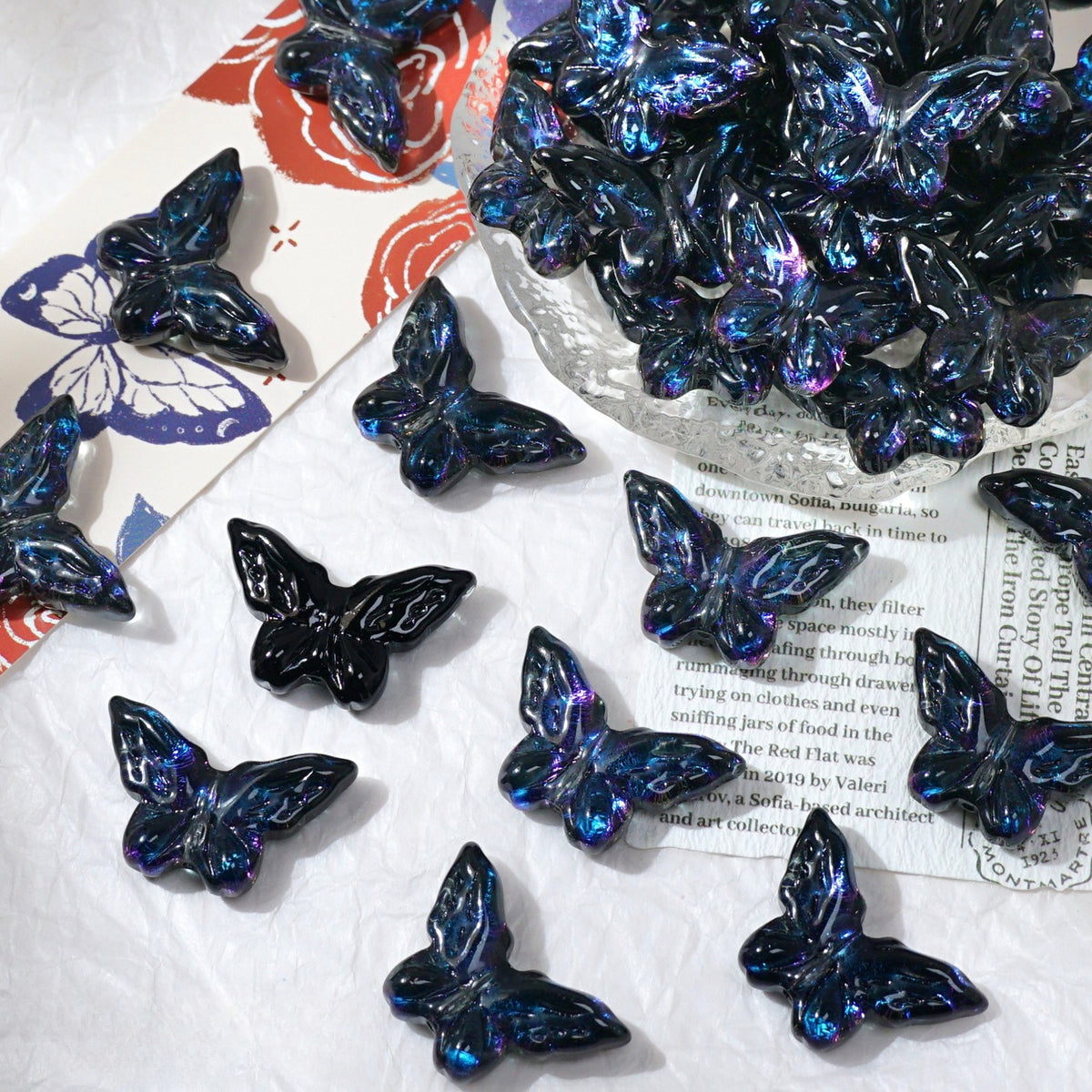 Multiple glossy iridescent butterfly-shaped beads scattered on a white textured surface