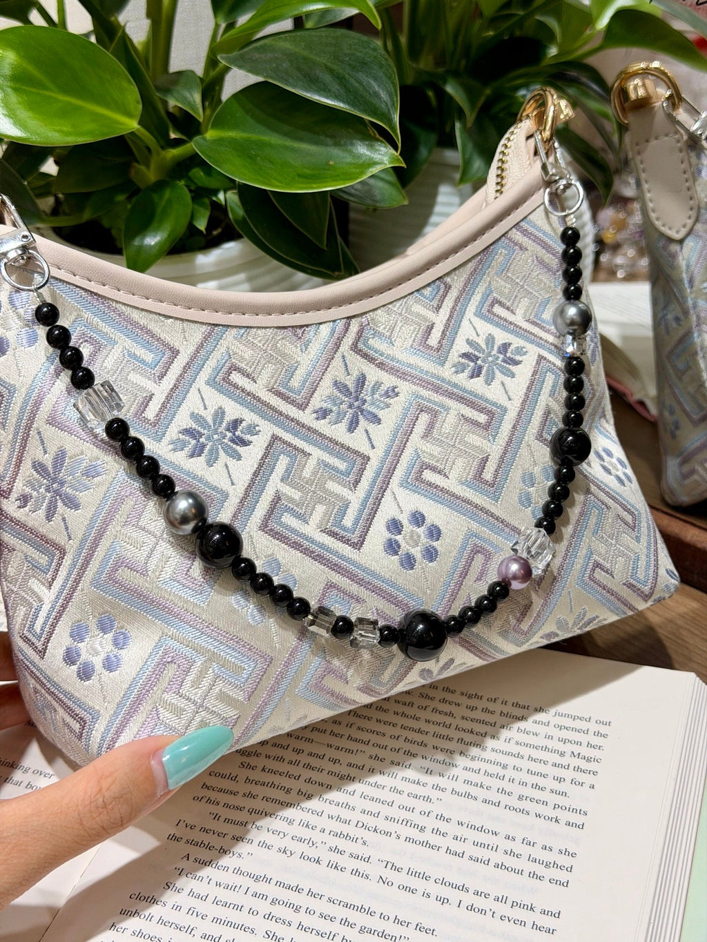 Beaded bag charm with black beads and clear crystals draped on a patterned shoulder bag over an open book.