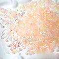 A pile of small translucent decorative beads in soft pastel pink, peach, pale yellow, and white, scattered on a white surface with printed script paper visible underneath.