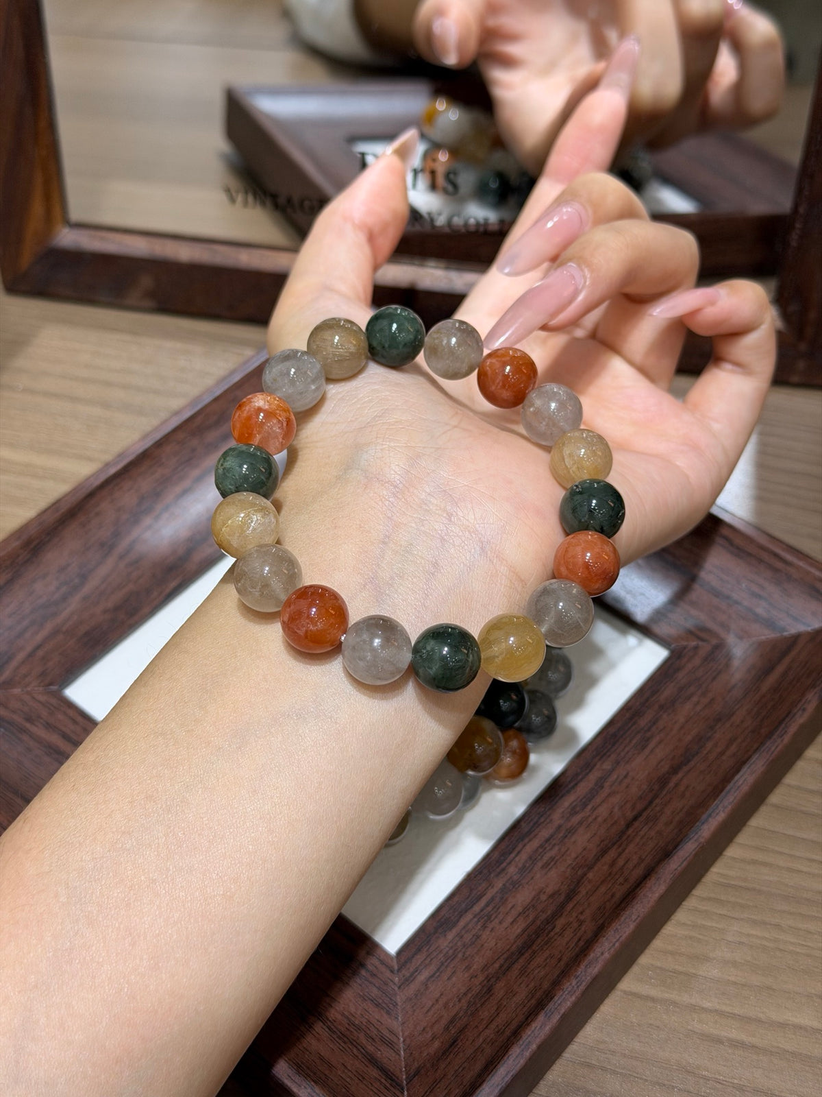 A hand holding a multicolor rabbit-hair quartz round-bead bracelet in shades of orange, green, gray, and milky beige, above a dark wooden tray