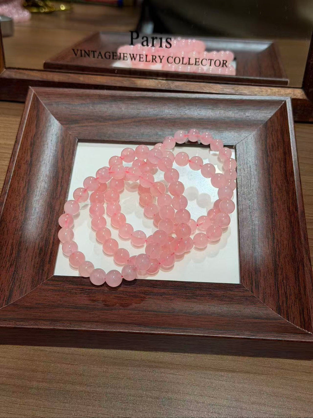 Pink translucent round beaded bracelet coiled in a dark brown wooden frame tray