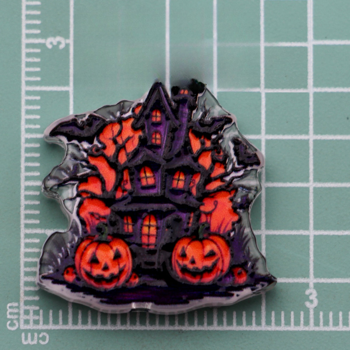 Halloween acrylic bead with a purple haunted house, jack-o'-lantern pumpkins, and bats on a cutting mat