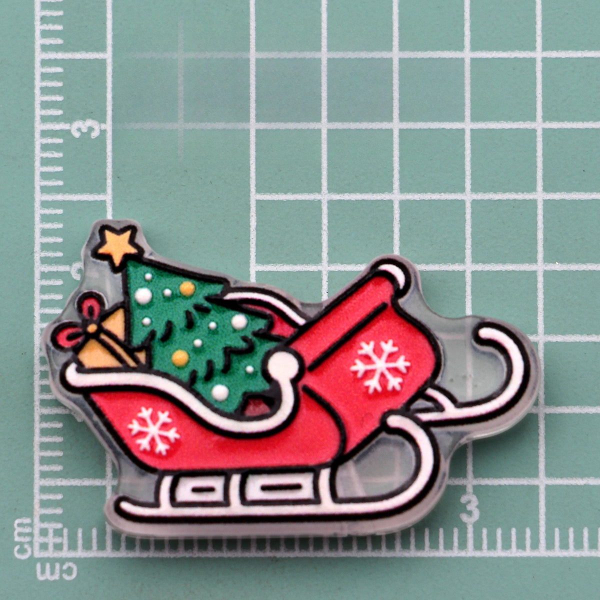 Red sleigh acrylic charm carrying Christmas tree and gift on green cutting mat