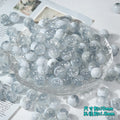 White and clear ice-crackle glass beads piled in a clear glass dish on a white surface with paper props
