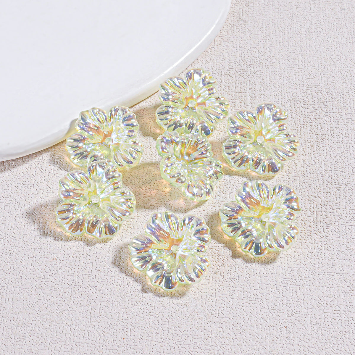 Seven iridescent resin flower beads arranged on a textured beige surface by a white plate edge