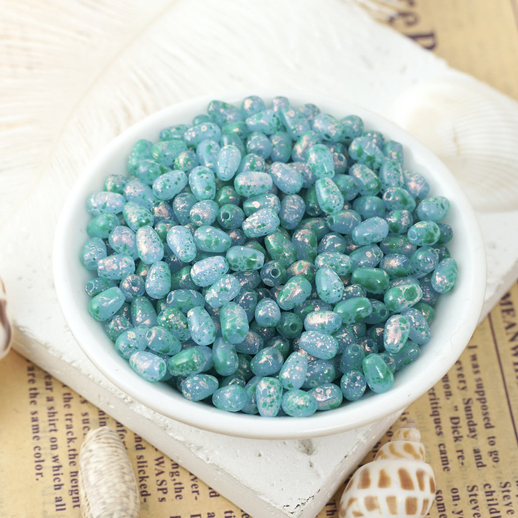 Iridescent blue and green speckled glass teardrop beads in a white ceramic bowl on vintage book pages with shells nearby.
