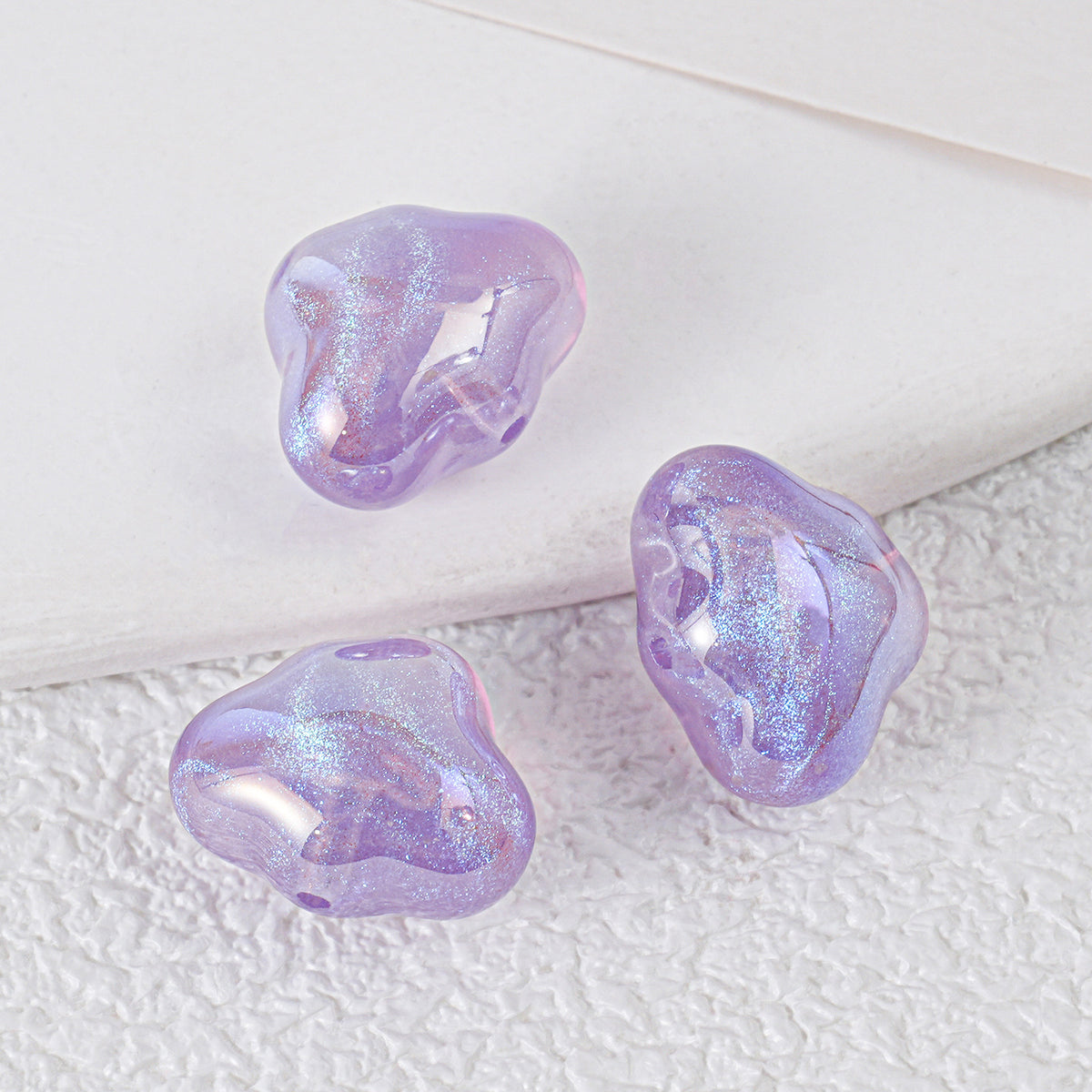 Three iridescent lavender-purple cloud-shaped resin beads with shimmering finish on a textured white surface
