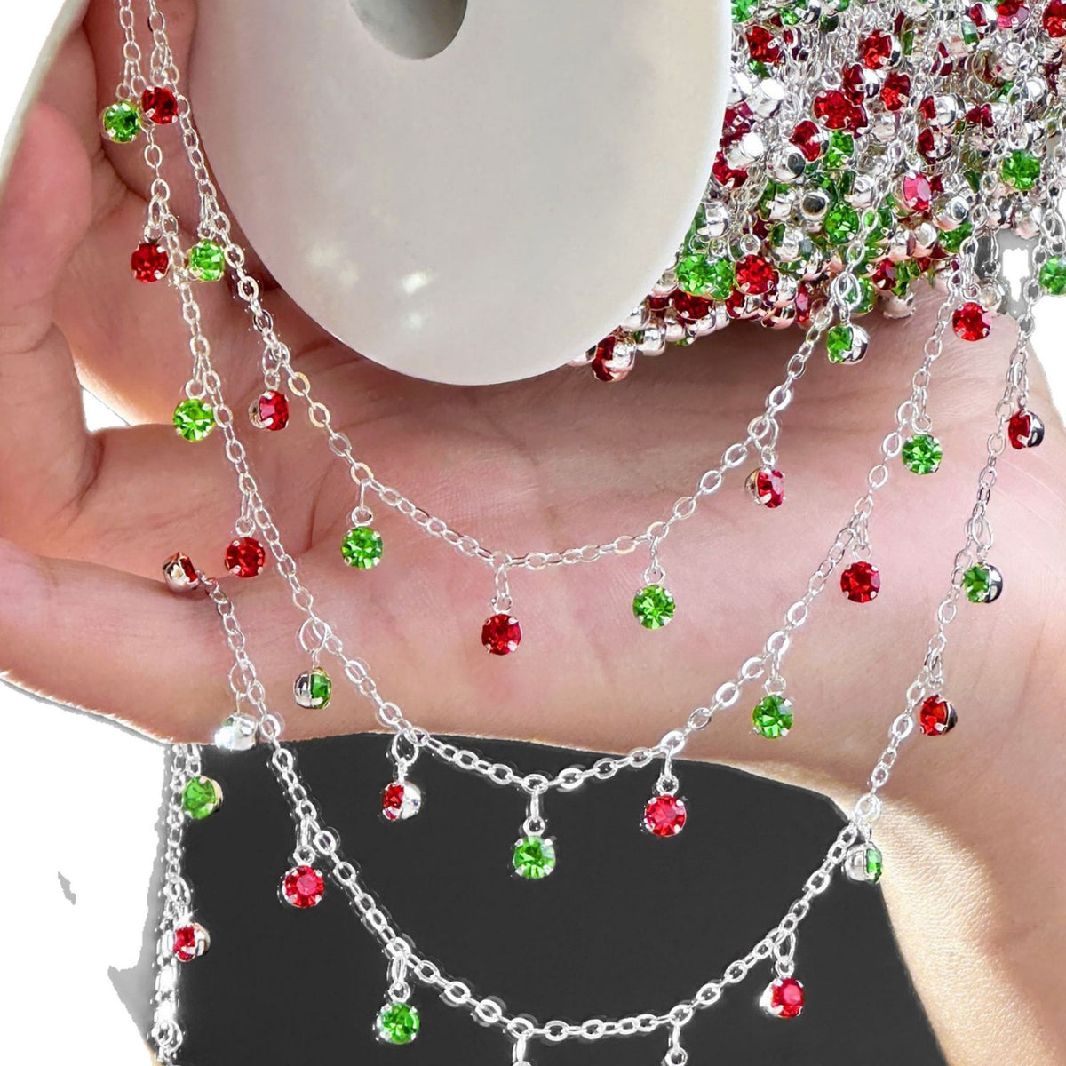A hand holding a spool of silver chain garlands decorated with small red and green faceted rhinestones, with more of the garland visible on the spool.