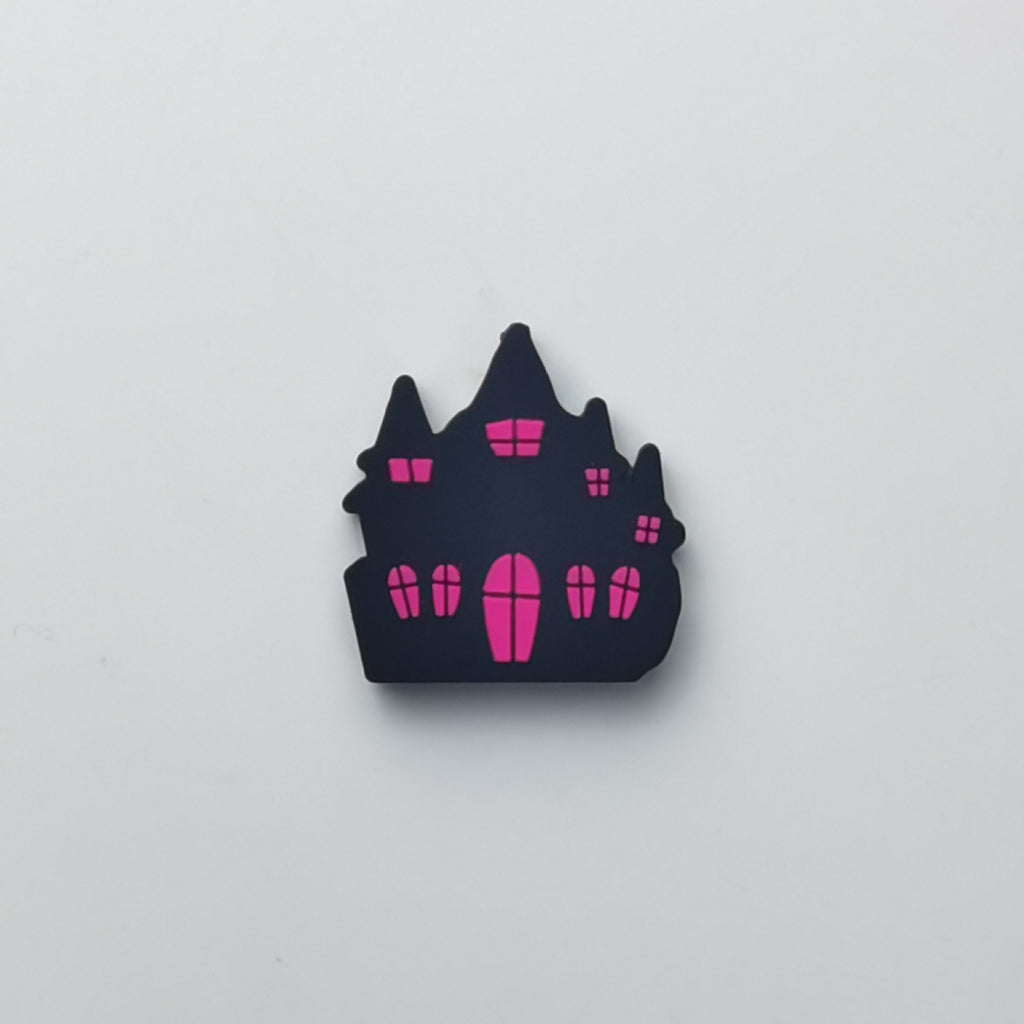 A small castle-shaped accessory, likely a pin, charm, or sticker, in matte black with bright pink windows and door details, centered on a plain light gray background.