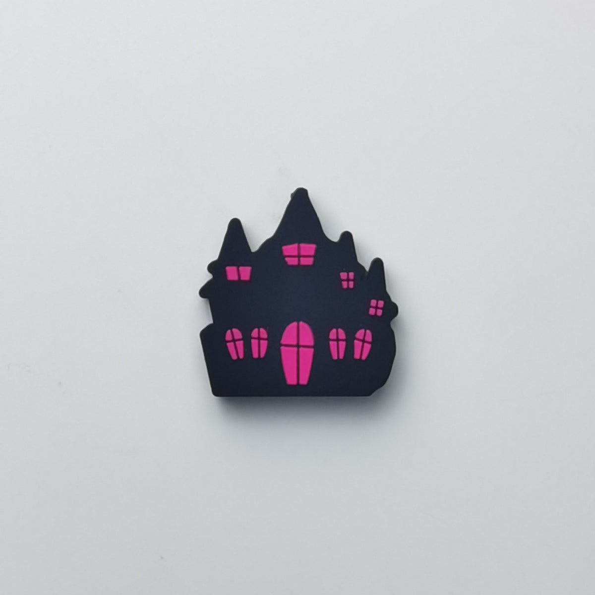 A small castle-shaped accessory, likely a pin, charm, or sticker, in matte black with bright pink windows and door details, centered on a plain light gray background.
